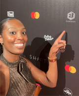 A person in a glittery dress beams as they point at the Eumelanin logo on a black backdrop adorned with various logos, including Mastercard. The setting seems to be a formal promotional event, highlighting Eumelanins allure and inviting guests to explore their exclusive shop.
