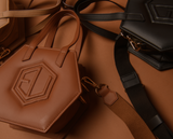 Hexagonal HEX Mini Bags in various shades of brown and black are spread out on a brown surface. The smaller versions feature stitched geometric patterns and have straps attached. The scene is warmly lit, highlighting the eumelanin-inspired tones.