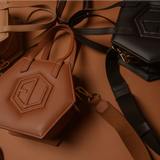 Hexagonal HEX Mini Bags in various shades of brown and black are spread out on a brown surface. The smaller versions feature stitched geometric patterns and have straps attached. The scene is warmly lit, highlighting the eumelanin-inspired tones.