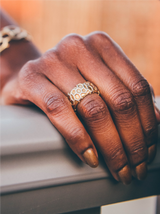 Close-up of a persons hand resting on a gray surface, adorned with a patterned gold ring and metallic gold nail polish. The background is softly blurred, highlighting the hands details—ideal inspiration for your next eumelanin-inspired beauty shop visit.