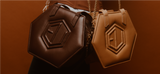 Two hexagonal leather bags, the HEX Mini Bag and its tan counterpart, feature embossed hexagon designs. Both showcase gold chains against a warm brown backdrop, their hues reminiscent of eumelanin tones - one dark brown and the other in light tan.