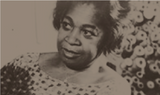 A sepia-toned image of a woman from Eumelanin Shop gazing off to the side. She is wearing a patterned dress and has short, curled hair. The background features a blurred floral design, enhancing the vintage feel of the photograph.