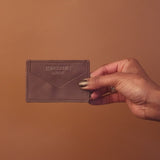 A hand holds a black leather wallet against a solid brown backdrop. The wallet, perfect for any shop enthusiast, features embossed text and a minimalist design that echoes the classic hues of eumelanin.