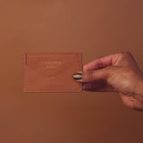 A hand elegantly showcases a tan leather cardholder against a matching brown background, with Eumelanin Detroit embossed on its surface. The persons nails gleam in gold, perfectly complementing this exquisite piece available for purchase at the Eumelanin shop.