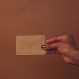A hand holds a small tan leather card holder against a matching brown background, showcasing its eumelanin-inspired hue. The holder features intricate stitching details and embossed text at the top, perfect for the discerning shop-goer.