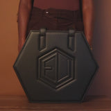 A black, hexagon-shaped leather handbag with two handles set against a brown backdrop. Featuring embossed initials AJ at the center, this HEX Bag boasts premium hardware that enhances its sophisticated allure.