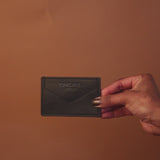 A hand, nails painted dark with the richness of eumelanin, holds a dark brown leather wallet against a solid brown backdrop. The wallet is embossed with text—a perfect accessory to shop in style.