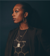 A woman with long, dark hair rich in eumelanin is gazing to the side against a dark background. She wears geometric earrings, a blazer, and a shirt featuring a hexagonal chemical structure design. Her layered necklaces shimmer subtly, each piece perfectly curated as if from an exclusive shop.