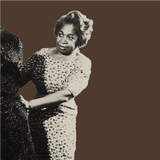 Historic photograph of a woman with short hair wearing a patterned dress, adjusting fabric on a mannequin in an elegant shop. The image, rich in eumelanin contrasts, is black and white against a solid brown background.