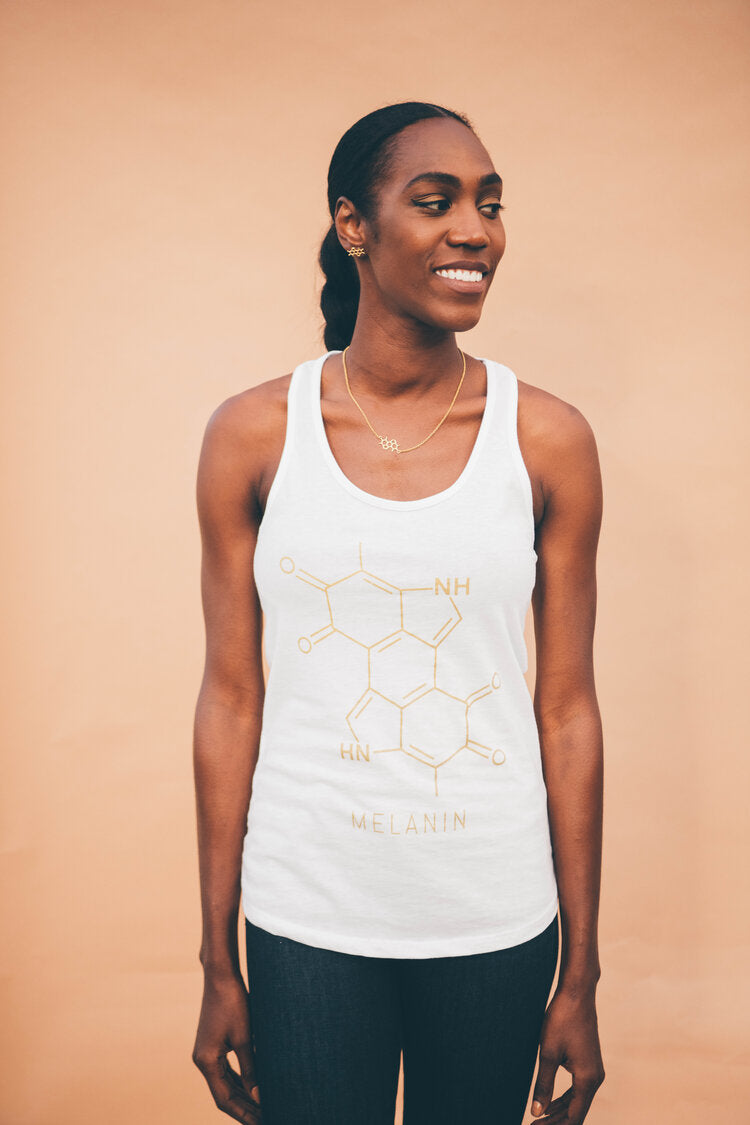 A person wearing a womens tank featuring a melanin chemical structure stands smiling against a peach-colored background. Their dark hair is tied back, and they wear a necklace, perfectly complementing the intricate design on their top.