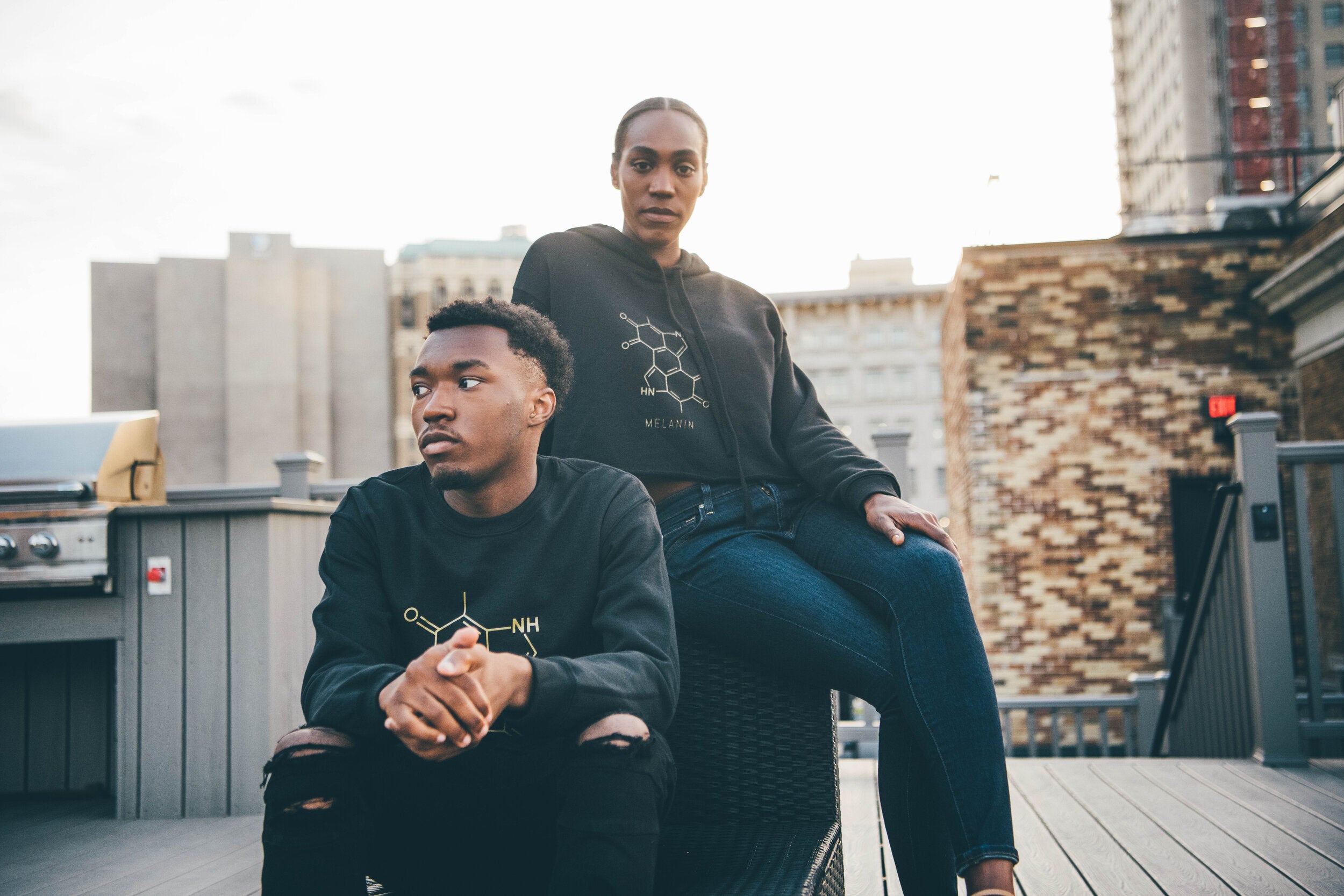A man and woman sit on a rooftop deck in an urban setting. Both wear black sweatshirts with chemical structure designs in gold print. The man sits on the edge of a bench with hands clasped, while the woman, in a black crop hoodie, sits behind him with one hand on the bench.
