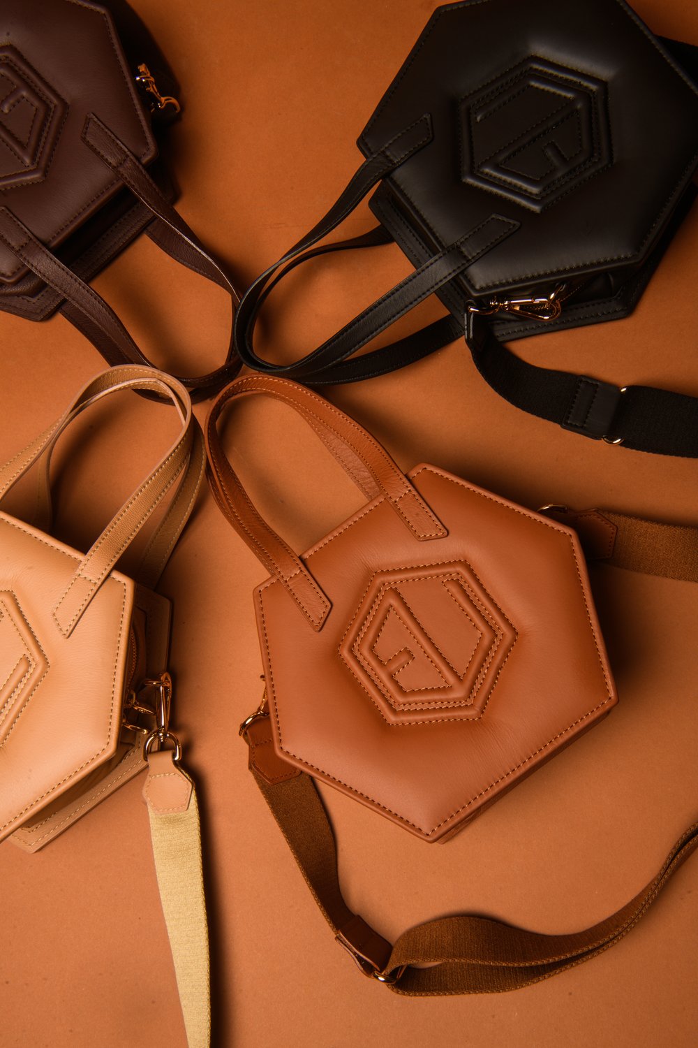 Four hexagonal-shaped handbags in black, brown, and beige are artfully arranged on an orange background. Each bag, available at Eumelanin Shop, boasts a short handle and strap with intricate geometric stitching on the front.